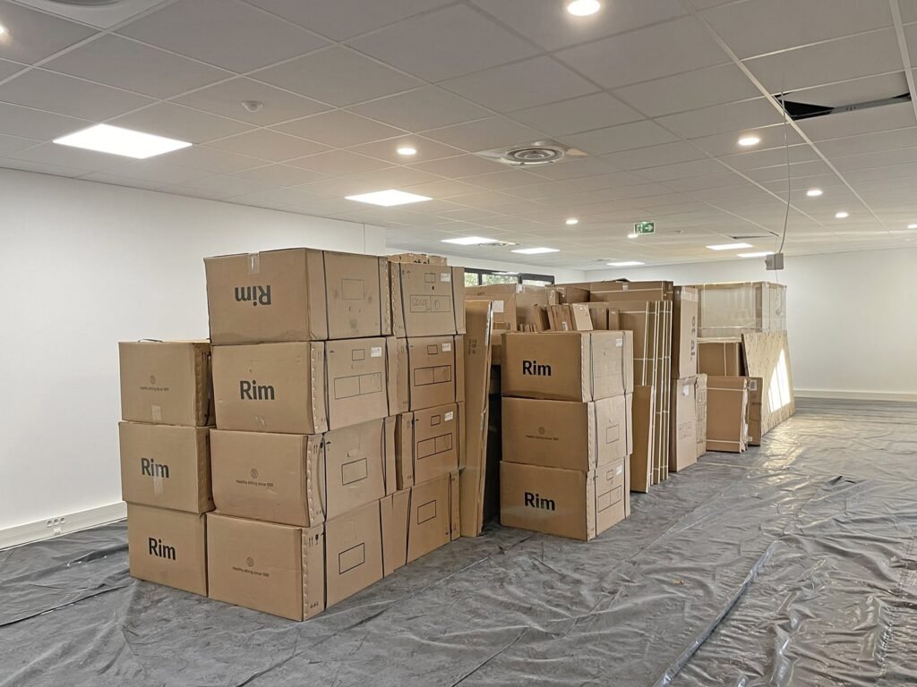 Une pile de grandes boîtes en carton étiquetées "Rim" est disposée au milieu d'une pièce lumineuse et non meublée, dont le sol est recouvert d'une feuille de plastique.