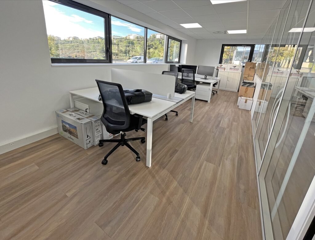 Bureau moderne avec plusieurs bureaux vides, des chaises de bureau, du matériel informatique et des boîtes, avec de grandes fenêtres et un sol en bois.