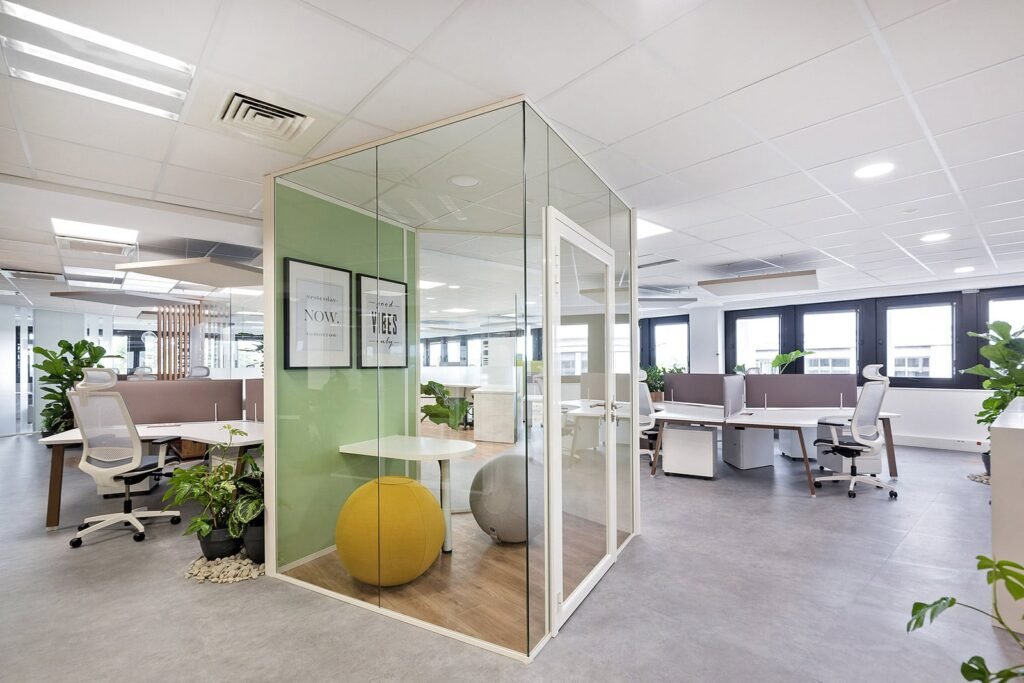 Bureau moderne avec salle de réunion vitrée, chaises ergonomiques, bureaux, plantes en pot et grandes fenêtres offrant une lumière naturelle.