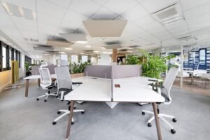 Bureau moderne à aire ouverte avec des bureaux blancs, des chaises ergonomiques, des écrans de séparation et des plantes d'intérieur ; de grandes fenêtres apportent de la lumière naturelle.