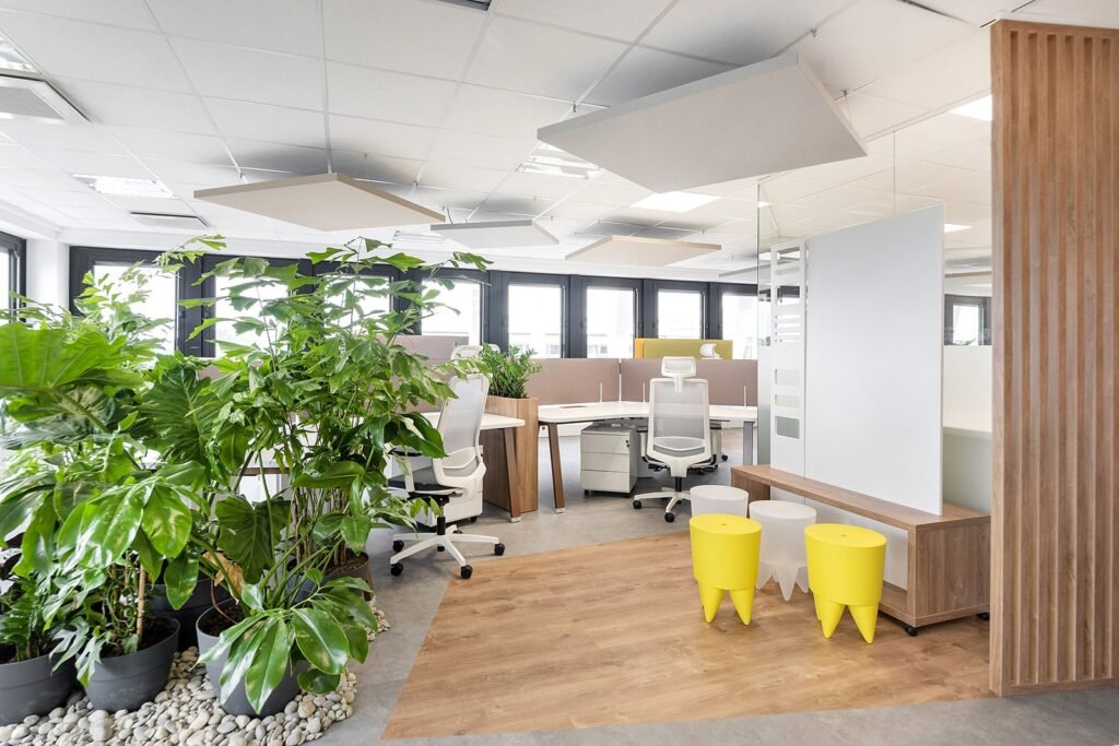 Espace de bureau moderne avec bureaux et chaises blancs, grandes plantes vertes et tabourets jaunes sur un sol en bois et en pierre. La lumière naturelle pénètre par de grandes fenêtres.