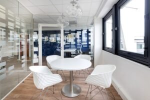 Une salle de réunion moderne avec une table ronde blanche, quatre chaises blanches, des murs en verre avec des motifs géométriques, un sol en bois et de grandes fenêtres.