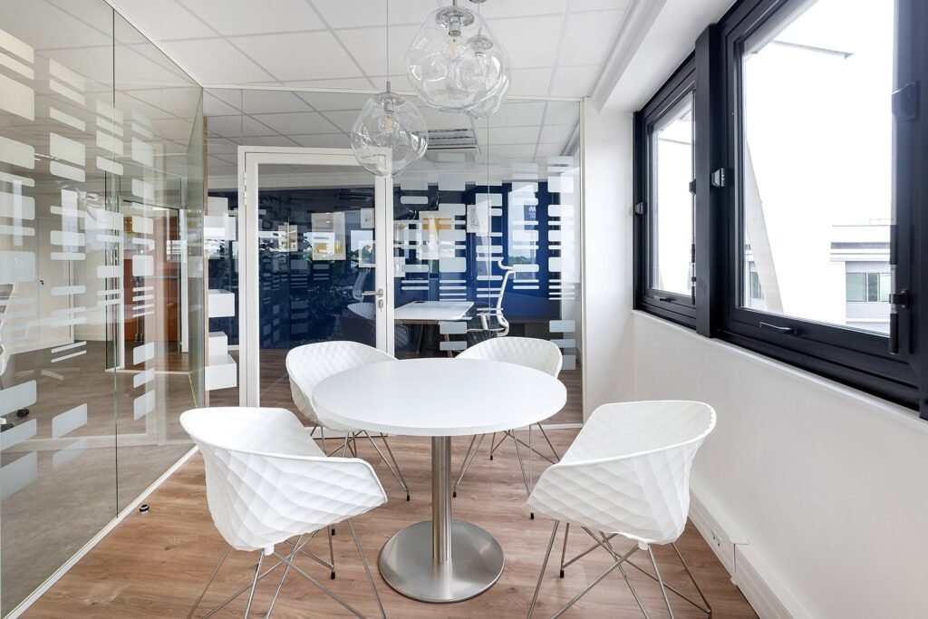Une salle de réunion de bureau moderne avec une table ronde blanche, quatre chaises blanches, des murs en verre avec des motifs géométriques et de grandes fenêtres laissant entrer la lumière naturelle.