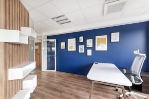 Bureau moderne avec un mur d'accent bleu, des œuvres d'art encadrées, un bureau blanc, une chaise ergonomique, un sol en bois et des étagères. Une porte vitrée mène à une autre pièce.