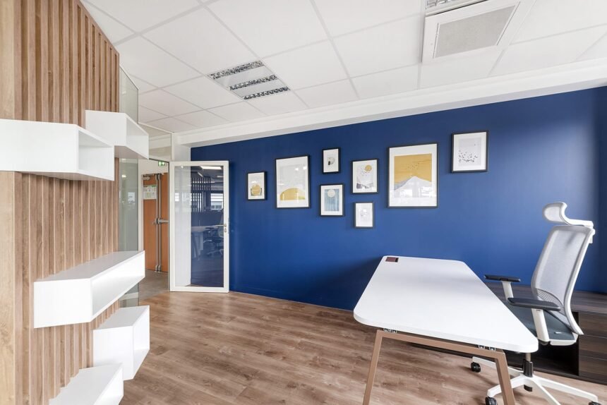Bureau moderne avec un mur d'accent bleu, des œuvres d'art encadrées, un bureau blanc, une chaise ergonomique, un sol en bois et des étagères. Une porte vitrée mène à une autre pièce.