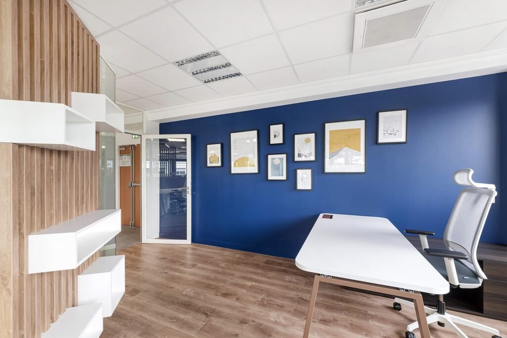 Bureau moderne avec un mur d'accent bleu, une œuvre d'art encadrée, un sol en bois, un bureau blanc et une chaise ergonomique. Des étagères et une porte vitrée sont visibles sur le côté gauche.