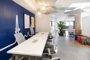 Un espace de bureau moderne avec des bureaux blancs, des chaises ergonomiques, un mur d'accent bleu, des lampes suspendues, des plantes d'intérieur et une variété de sièges.