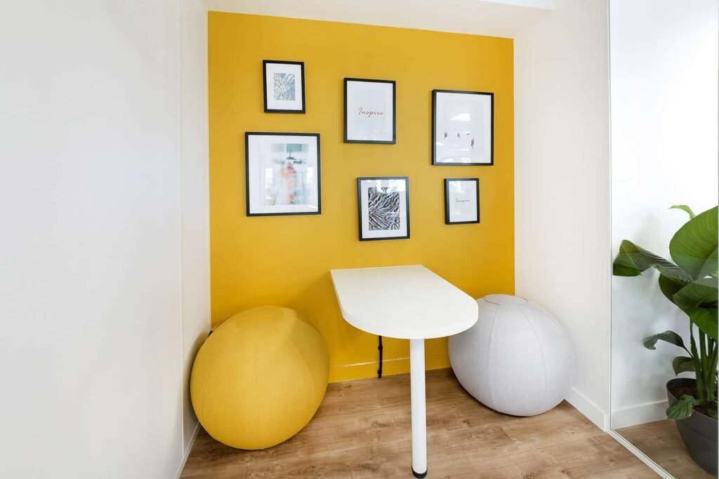 Un petit espace de travail comporte un bureau blanc, un ballon d'exercice jaune et un gris, un mur d'accent jaune avec des œuvres d'art encadrées et une plante en pot dans le coin.