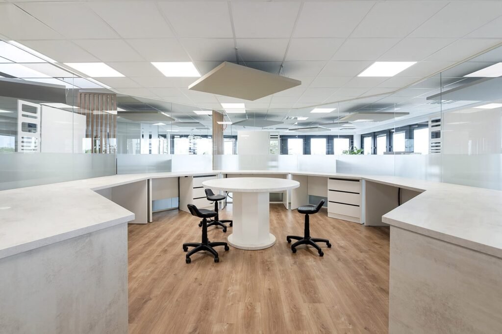 Bureau moderne avec une table ronde blanche, trois chaises pivotantes noires, un sol en bois et des cloisons en verre dépoli sous des plafonniers lumineux.