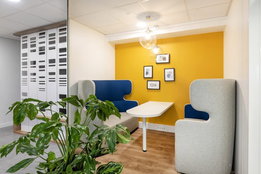 Un coin réunion de bureau moderne avec deux sièges gris et bleus, une table blanche, un mur d'accent jaune, trois tableaux encadrés, des lampes suspendues et une plante verte au premier plan.