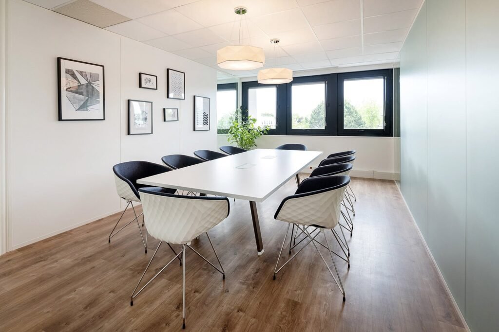 Une salle de conférence moderne avec une table rectangulaire blanche, huit chaises, des œuvres d'art murales, de grandes fenêtres et une plante sur un sol en bois.