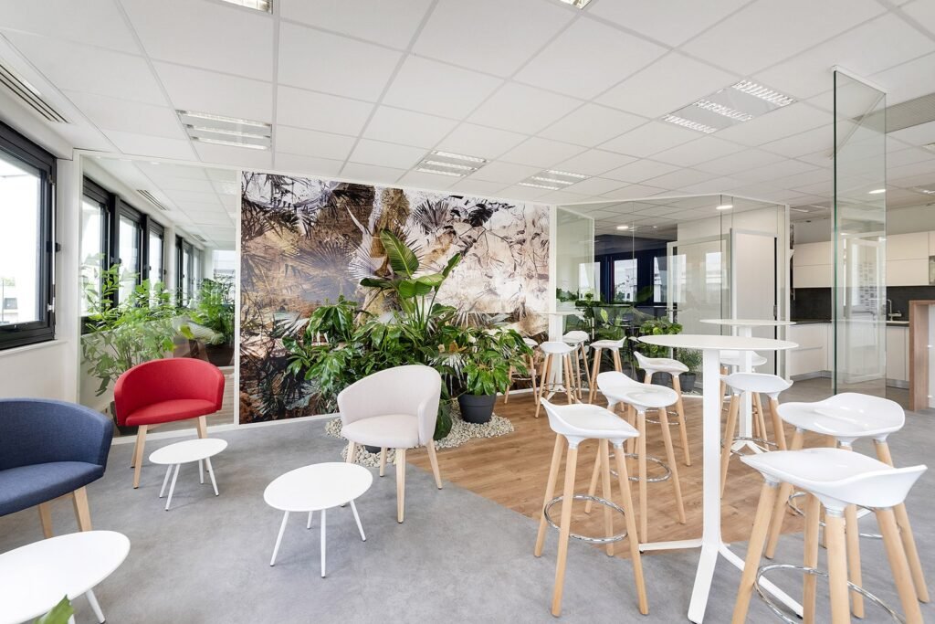 Salon de bureau moderne avec des tables et des chaises blanches, des fauteuils, des plantes et une peinture murale sur le thème de la nature. De grandes fenêtres apportent la lumière naturelle ; des cloisons en verre séparent les espaces.