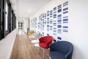 Un couloir avec un sol en bois, des chaises modernes de différentes couleurs, de petites tables rondes et un mur décoré d'un motif abstrait bleu.