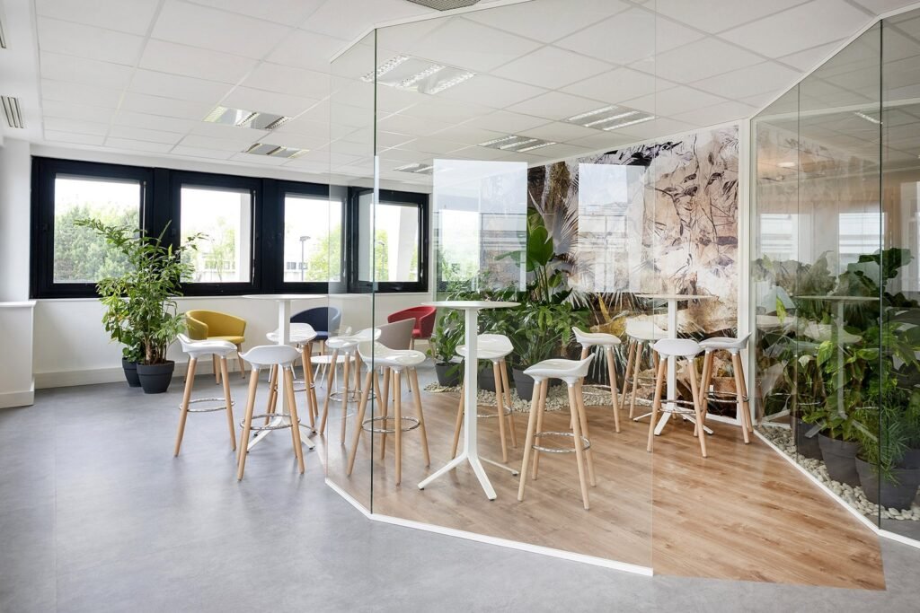 Salon de bureau moderne avec tables hautes, tabourets de bar blancs, cloisons en verre, plantes d'intérieur et grandes fenêtres laissant entrer la lumière naturelle.