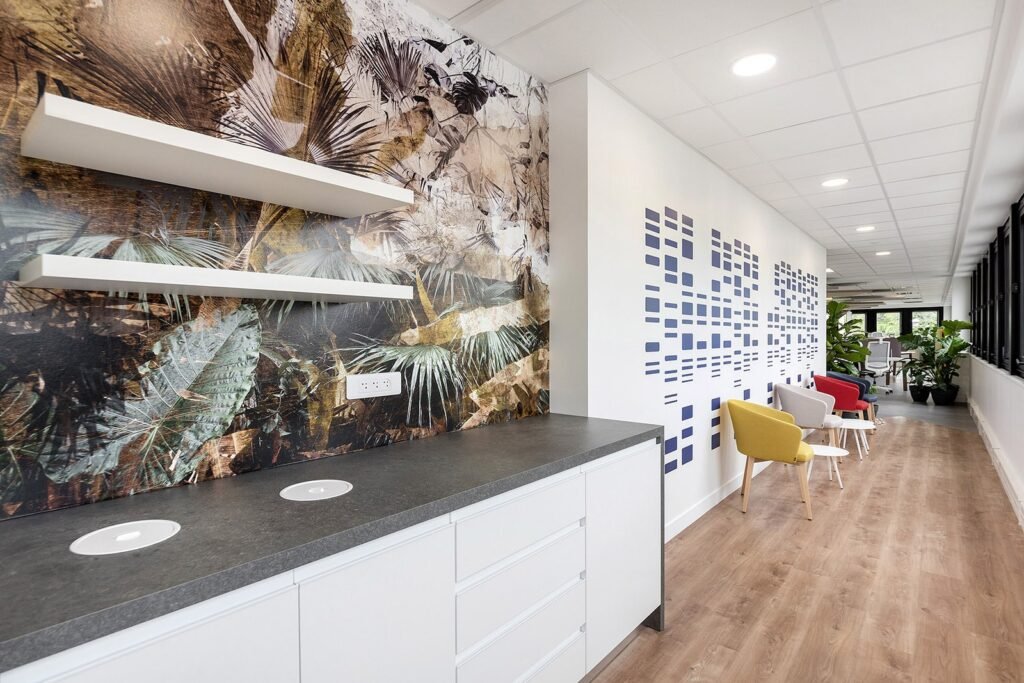 Couloir de bureau moderne avec une kitchenette, des œuvres d'art murales abstraites, des étagères et une rangée de chaises colorées sur le côté droit ; de grandes fenêtres apportent de la lumière naturelle.