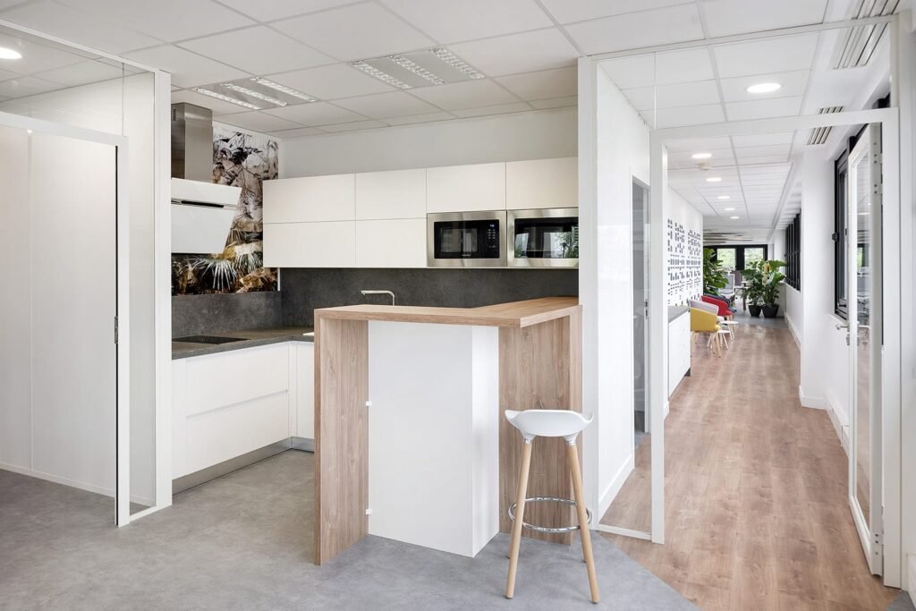 Kitchenette de bureau moderne avec des armoires blanches, un micro-ondes intégré, un petit comptoir de bar en bois et un tabouret, adjacente à un couloir avec un sol en bois et des murs en verre.