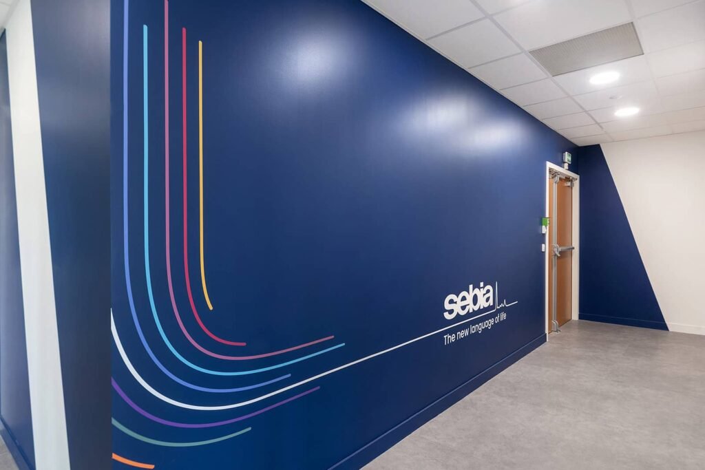 Un couloir aux murs bleus avec des lignes courbes colorées et le logo Sebia à côté d'une porte en bois fermée ; le plafond est blanc avec des lumières encastrées.
