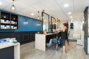 Un salon de coiffure moderne avec un mur d'accent bleu, des étagères de produits capillaires et un styliste qui coupe les cheveux d'une cliente à un poste de coiffure.