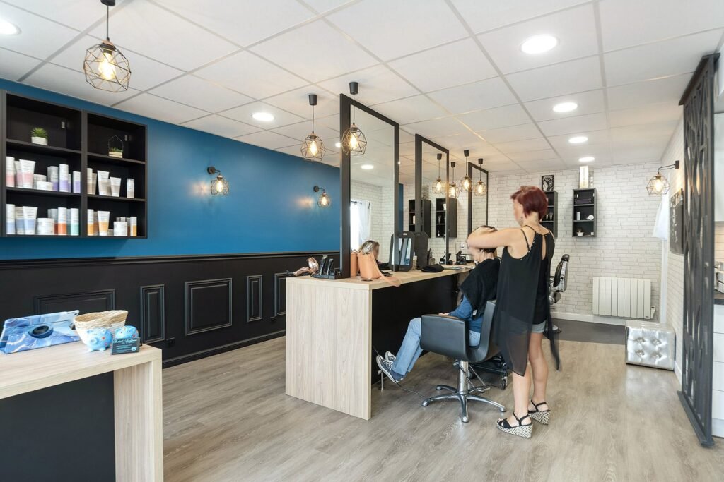 Un coiffeur coupe les cheveux d'une cliente dans un salon moderne aux murs bleus et blancs, au sol en bois et aux étagères présentant des produits capillaires.