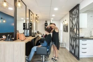 Un coiffeur coupe les cheveux d'une cliente assise dans un salon moderne et bien éclairé, avec des miroirs, des fauteuils de salon et des produits de beauté visibles.