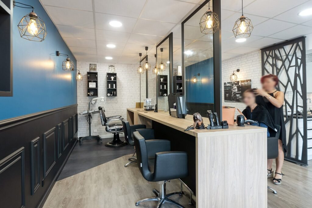 Intérieur d'un salon de coiffure moderne avec plusieurs stations de coiffage, des chaises de salon noires et un styliste travaillant avec un client. Le décor comprend des murs bleus et blancs et des lampes suspendues.