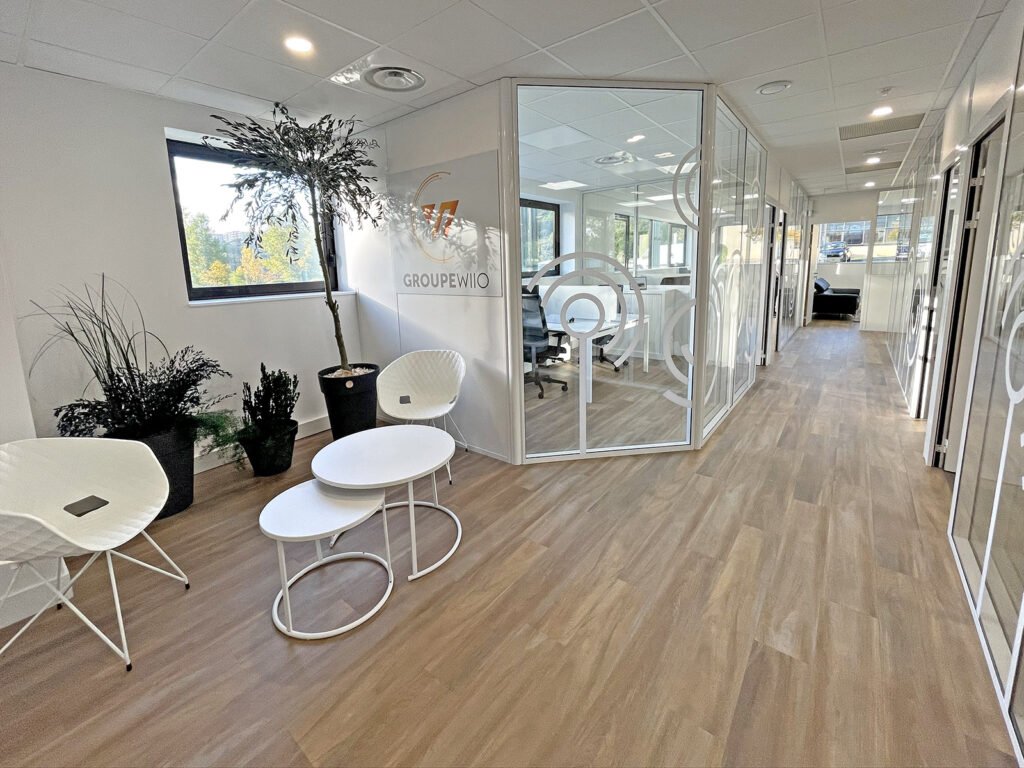 Couloir de bureau moderne avec sol en bois, salles de réunion vitrées, chaises blanches, petites tables rondes et plantes en pot. La lumière naturelle pénètre par les fenêtres.