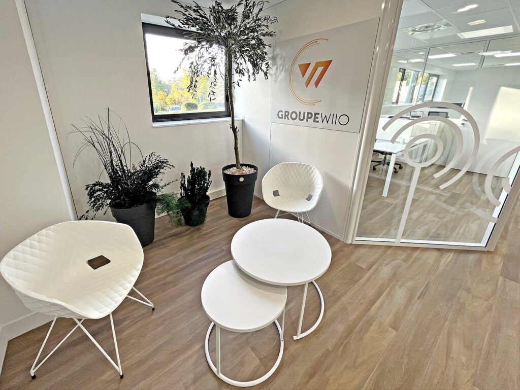 Salle d'attente d'un bureau moderne avec deux chaises blanches, des tables rondes emboîtées, des plantes en pot et une salle de réunion vitrée ; logo "GROUPE WIIIO" sur le mur.