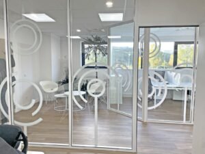 Intérieur de bureau moderne avec des cloisons en verre présentant des motifs circulaires, des chaises blanches, des petites tables et de grandes fenêtres laissant entrer la lumière naturelle.