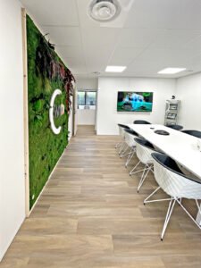 Salle de conférence moderne avec une longue table blanche, des chaises noires et blanches, un mur de mousse verte avec les lettres "CWD" et un écran mural affichant une image de chute d'eau.