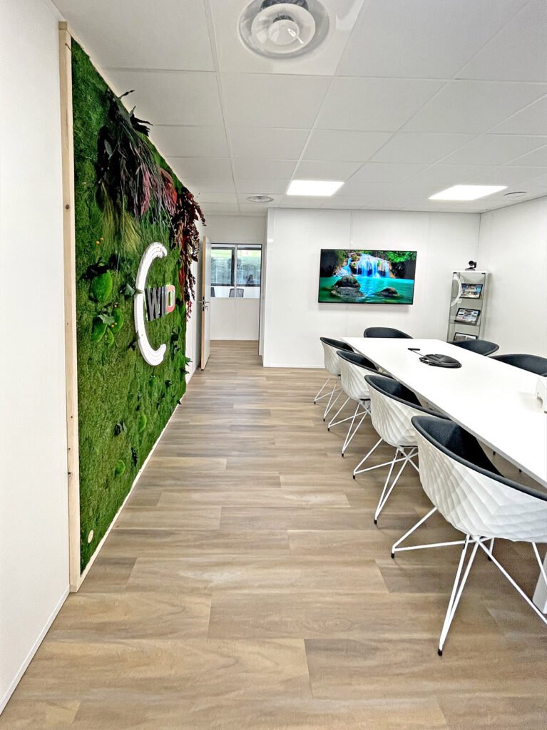 Salle de conférence moderne avec une longue table, des chaises noires et blanches, un mur de plantes vivantes vertes et un téléviseur mural affichant une image de cascade.