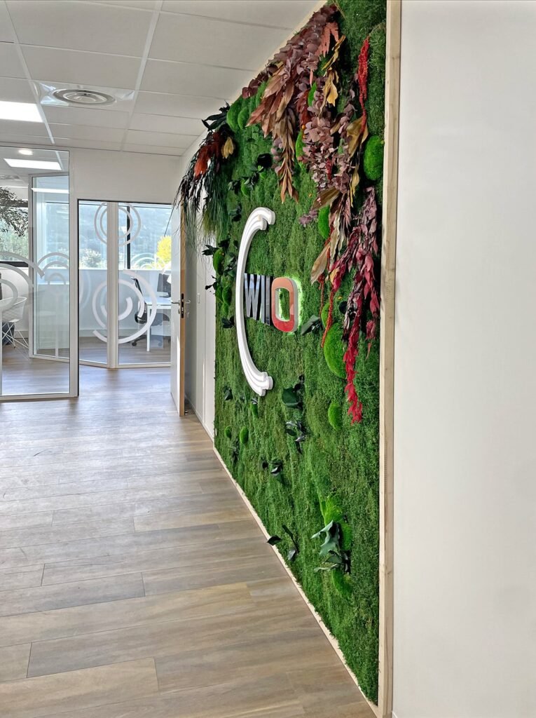 Dans un couloir au sol en bois, un mur en mousse verte est décoré de feuillages et du logo de l'entreprise "WILO". Les murs des bureaux en verre sont visibles sur la gauche.