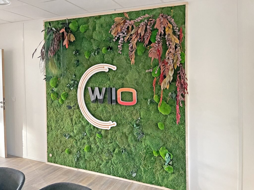 Un logo WIO est affiché sur un mur de mousse avec des fleurs et des plantes séchées.
