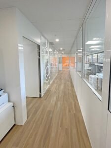 Un couloir de bureau moderne avec un sol en bois, des bureaux aux parois vitrées à gauche, et des boîtes près des fenêtres à droite. Le verre présente des motifs circulaires blancs.