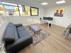 Un bureau moderne avec un canapé noir, un tapis à motifs, une table basse en bois, deux bureaux avec des ordinateurs, une grande fenêtre et une peinture murale.