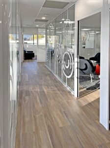 Un couloir de bureau moderne avec un sol en bois, des murs en verre avec des motifs circulaires et un coin salon avec des chaises noires visibles à travers une porte ouverte.