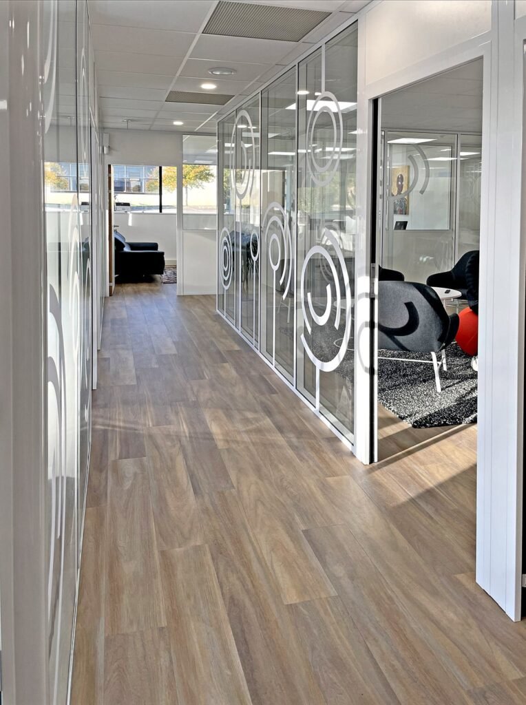 Un couloir de bureau moderne avec un sol en bois, des murs en verre avec des motifs circulaires et un coin salon avec des chaises noires visibles à travers une porte ouverte.