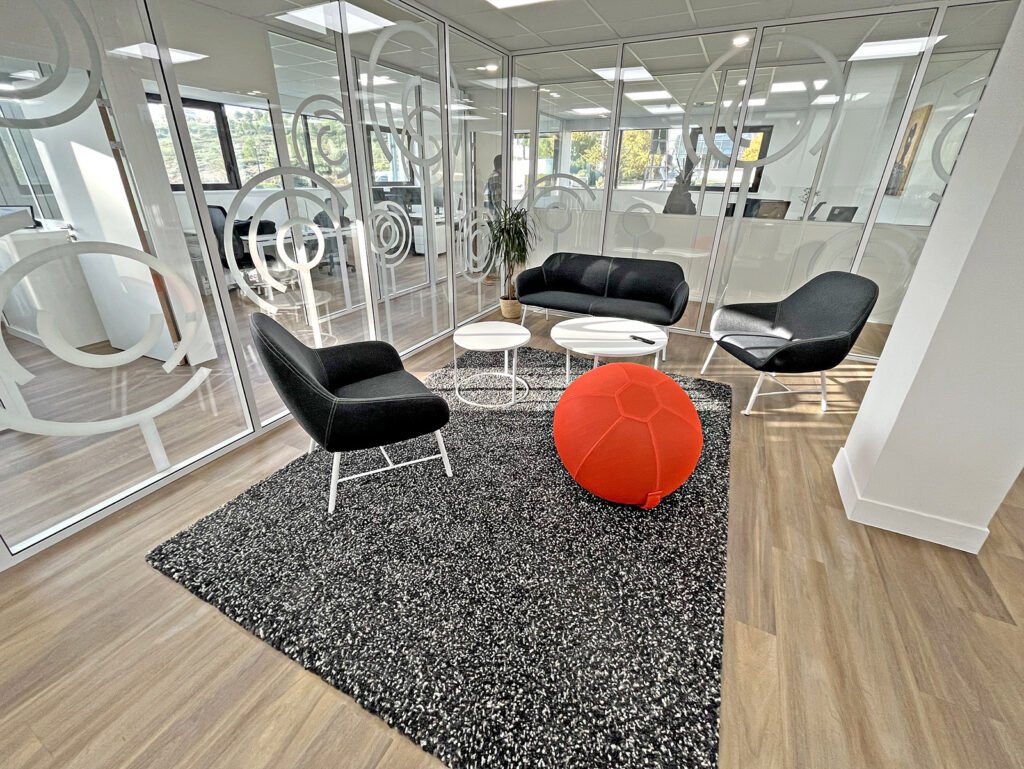 Salon de bureau moderne avec des chaises noires, un siège rouge en forme de boule, trois tables rondes blanches et un tapis sombre sur un sol en bois clair, entouré de murs en verre avec des motifs circulaires.