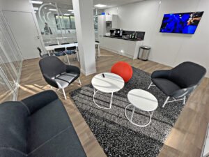 Salon de bureau moderne avec des chaises noires, un canapé, des tables rondes, un ottoman rouge, une kitchenette, une télévision murale et une salle de réunion avec une table et des chaises visibles à travers des parois en verre.
