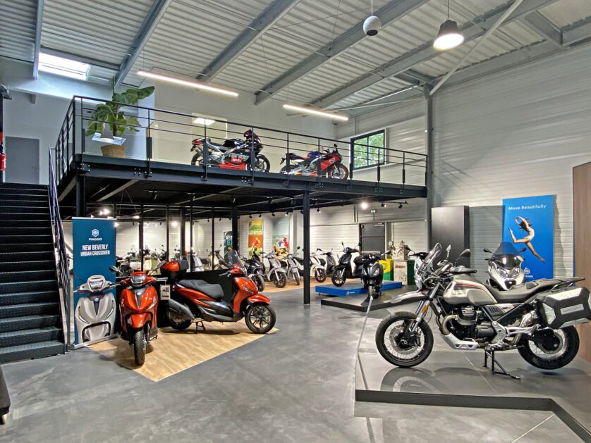 Une salle d'exposition de motos moderne avec différents modèles exposés, y compris des scooters et des motos de tourisme, au rez-de-chaussée et au niveau de la mezzanine.