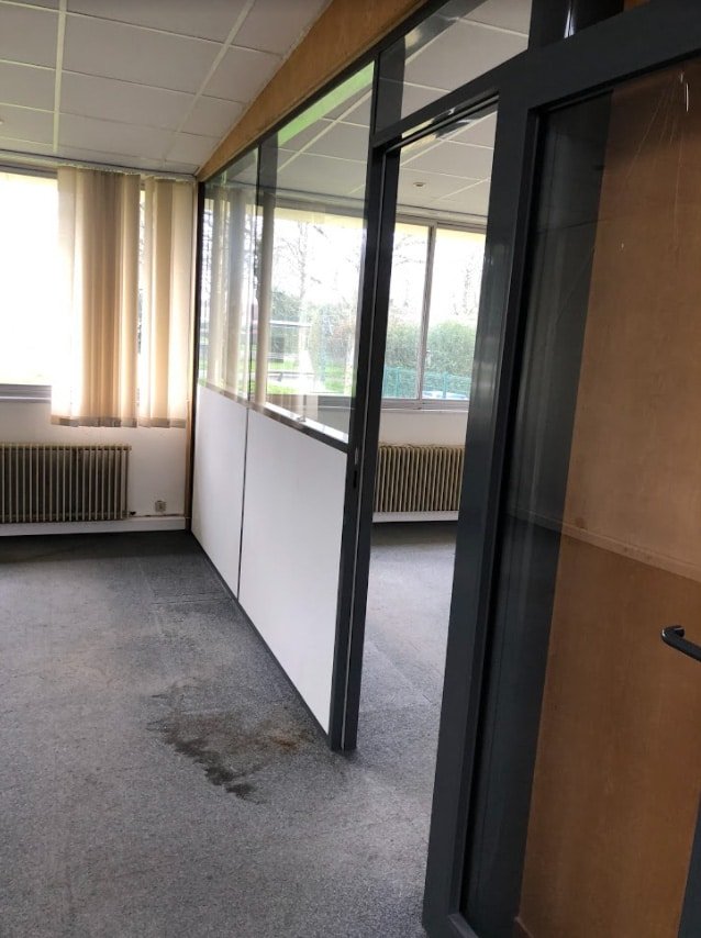 Un espace de bureau vide avec des cloisons en verre et en panneaux, un sol en moquette avec une tache visible, de grandes fenêtres et des stores verticaux.