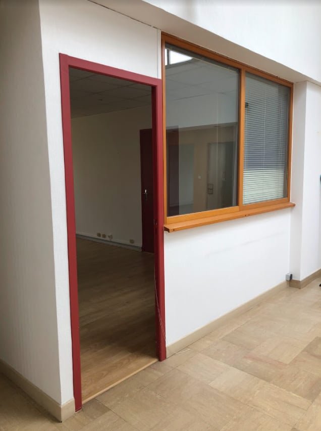 Une porte ouverte avec un cadre rouge mène à une pièce avec un sol en bois ; le mur adjacent comporte une fenêtre avec un cadre en bois et des stores.