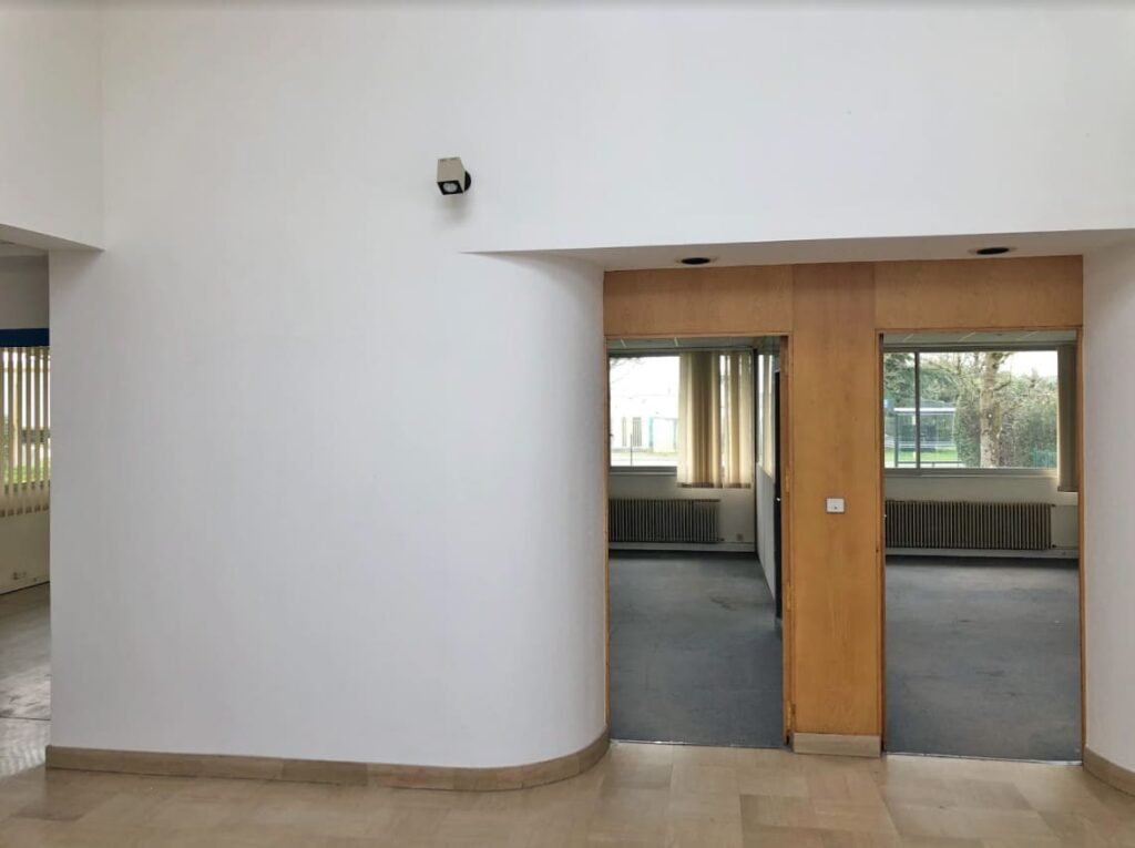 Une pièce aux murs blancs avec une double porte en bois mène à une autre pièce avec des sols recouverts de moquette et de grandes fenêtres montrant la verdure extérieure.