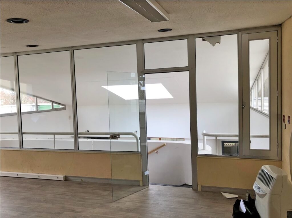 Espace de bureau avec un mur et une porte en verre, dont une vitre est cassée en haut à droite. Une cage d'escalier est visible au-delà de la vitre et un climatiseur portable se trouve dans le coin droit.