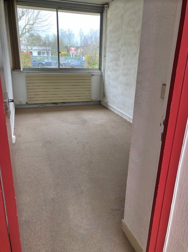 Petite pièce vide avec une moquette beige, des murs blancs et une grande fenêtre donnant sur un parking et des arbres, vue depuis une porte ouverte avec un cadre rouge.
