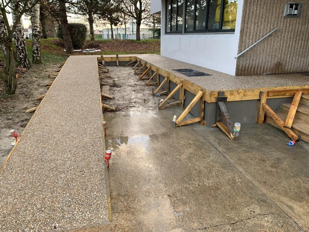 Un trottoir en béton est partiellement construit, avec des coffrages et des supports en bois en place le long de la section inachevée à côté d'un bâtiment.