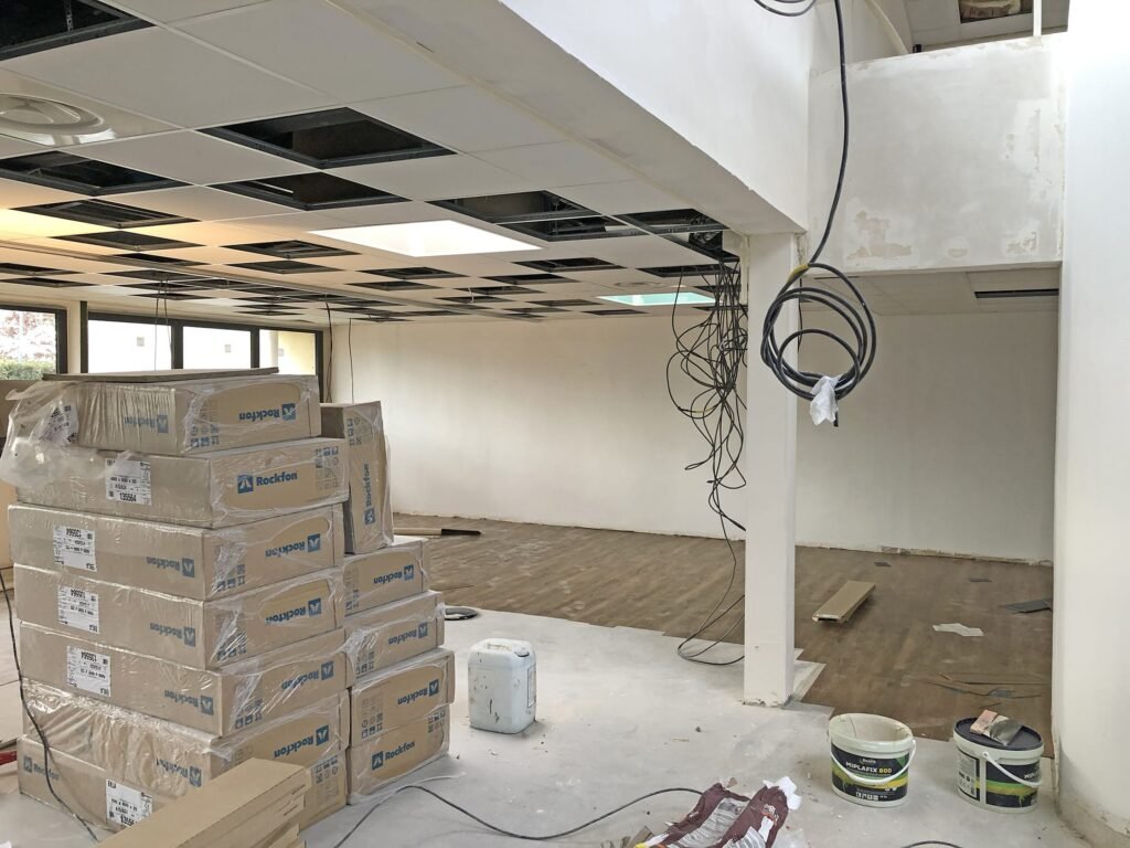 Une pièce en cours de rénovation avec des panneaux de plafond apparents, des fils suspendus, des boîtes empilées, des matériaux de construction et des outils éparpillés sur le sol.