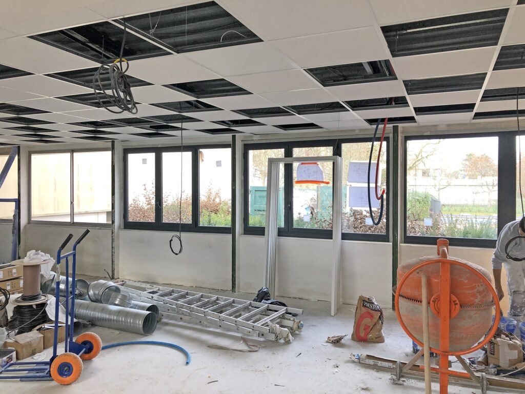 Espace de bureau en cours de construction avec câblage apparent, échelles, conduits de ventilation, outils et un plafond suspendu partiellement installé. De grandes fenêtres apportent de la lumière naturelle.