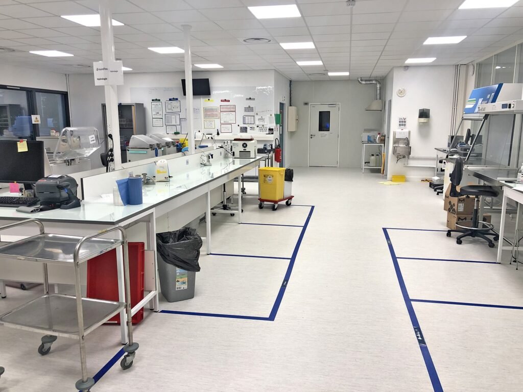 Un laboratoire propre et bien éclairé avec plusieurs postes de travail, du matériel de laboratoire, des poubelles et des marquages de sécurité au sol.