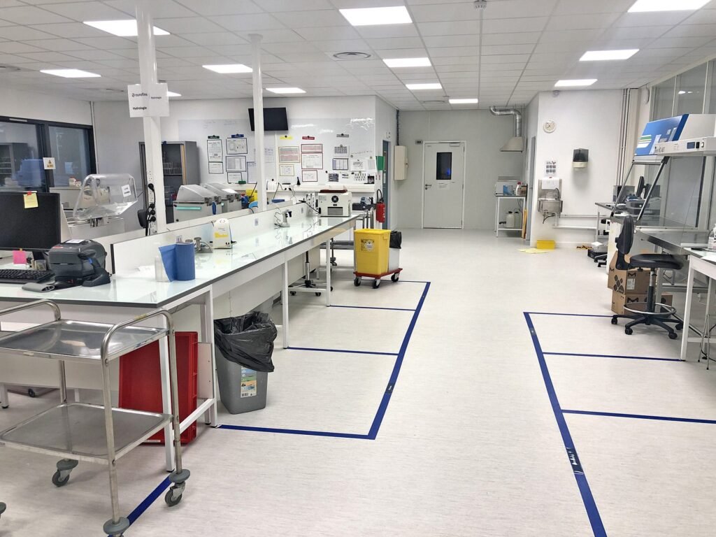 Laboratoire clair et stérile avec des établis, du matériel scientifique, des poubelles et des panneaux de sécurité. Du ruban bleu délimite des zones sur le sol. Aucune personne n'est présente.
