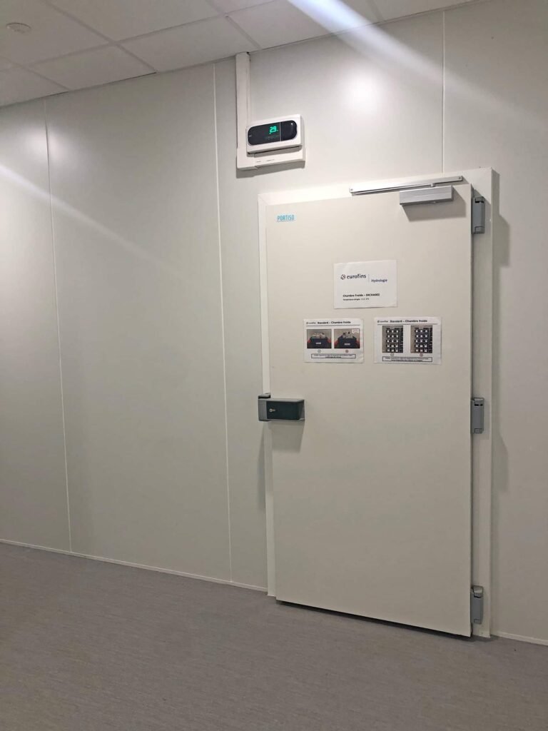 Une porte blanche, d'aspect industriel, dotée d'un clavier et de panneaux d'instructions, est encastrée dans un mur blanc ordinaire, sous un affichage numérique de la température.